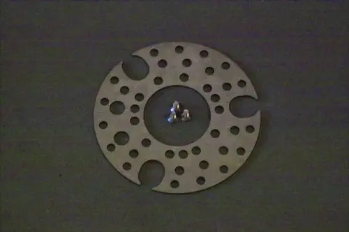 Brötje Stauscheibe mit mehreren Löchern und Einschnitten, in der Mitte angeordnet, umgeben von drei kleinen Metallkugeln auf dunklem Hintergrund.