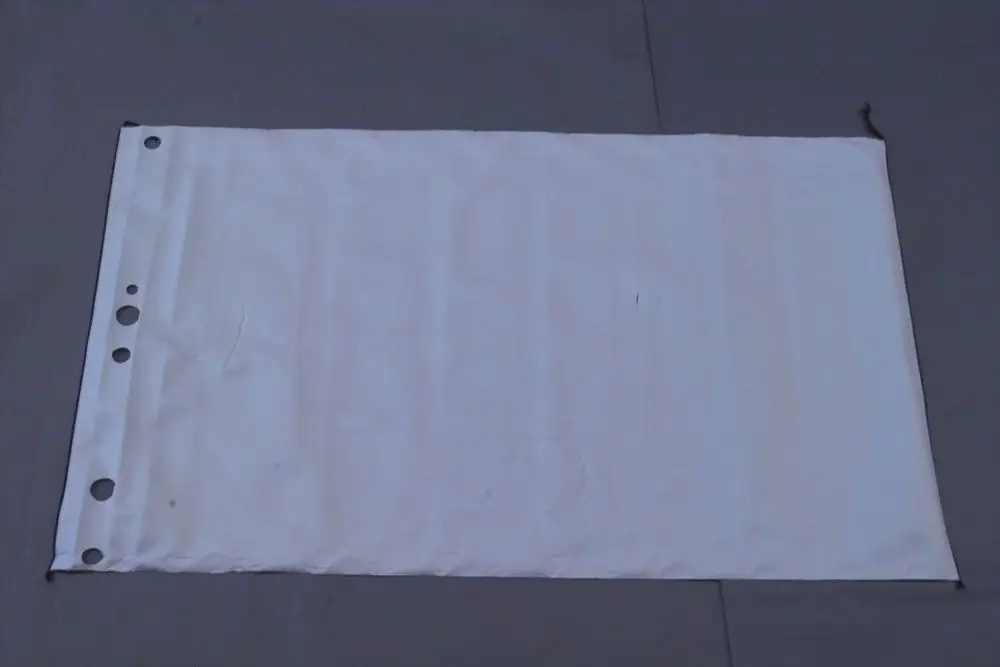 Weißes, rechteckiges Material mit Ösen an einer langen Seite, vermutlich zur Abdeckung oder Verkleidung, auf grauem Untergrund. Brötje ist als mögliche Marke erkennbar.