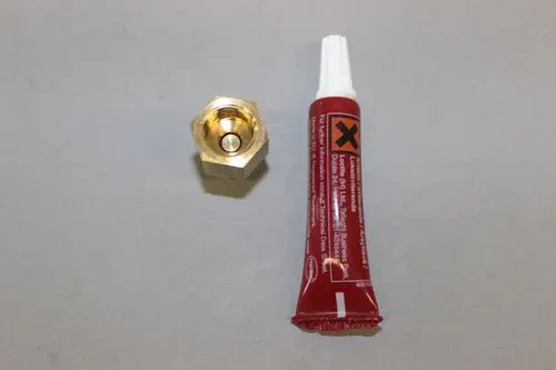 Goldfarbener Adapter für Druckanzeige, platziert neben einer roten Tube mit Kleber, auf grauem Hintergrund. Brötje ist der Herstellermarke zuzuordnen.