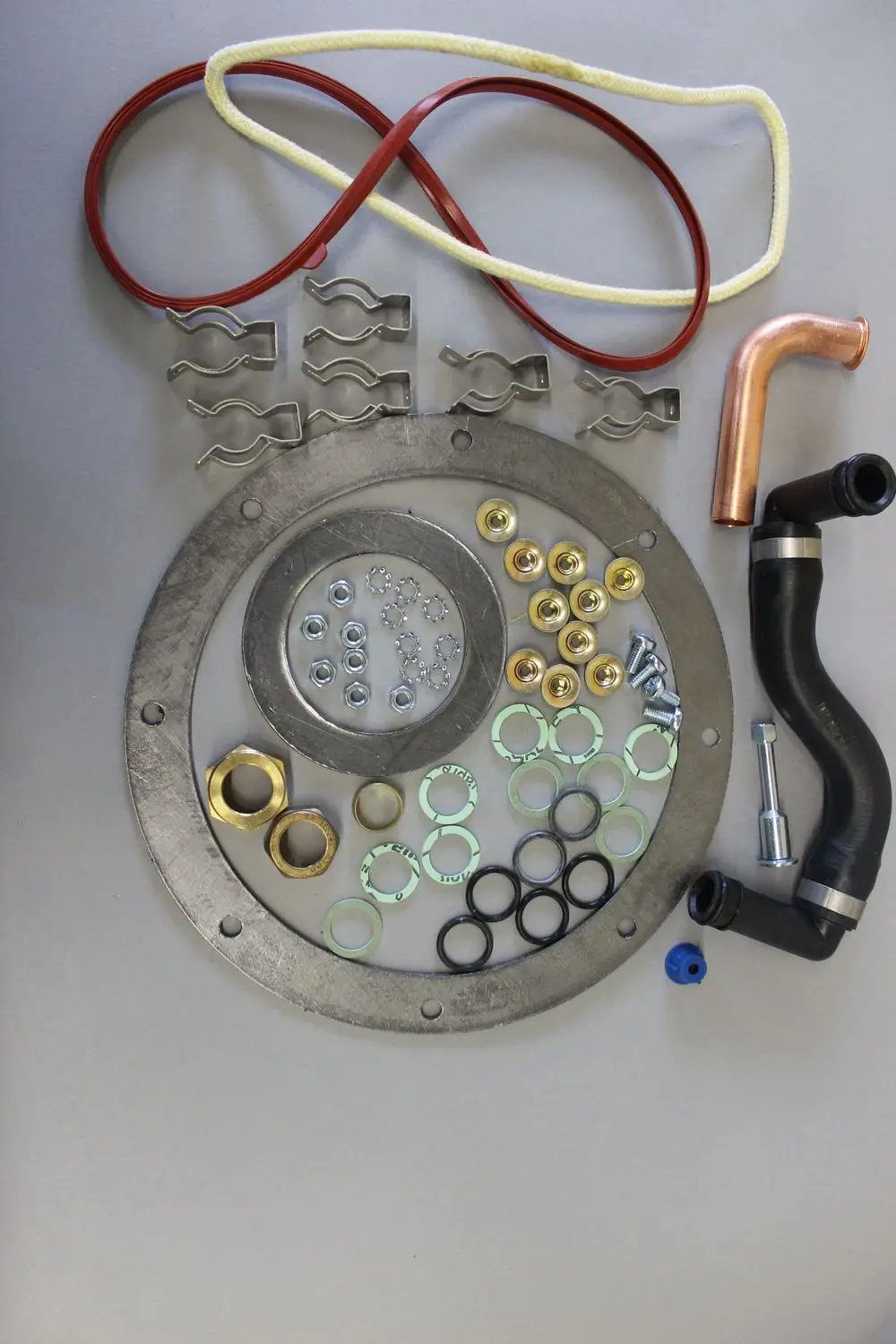 Brötje Zubehöre für einen Stirling Motor, darunter metallene Dichtungen, Schrauben, Riegel, Schläuche und Unterlegscheiben, auf grauem Untergrund angeordnet.