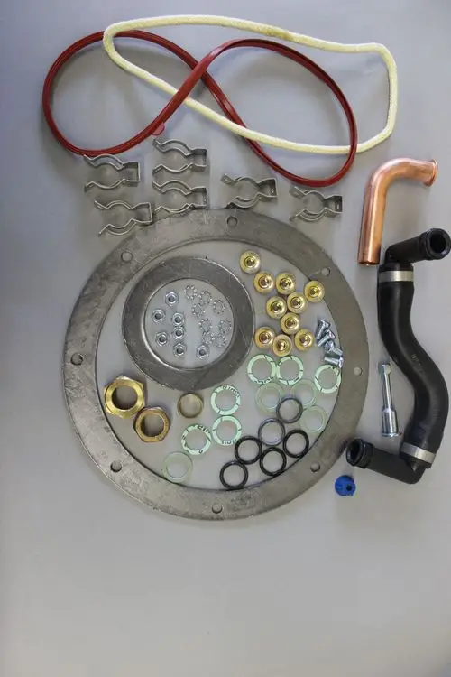 Brötje Zubehöre für einen Stirling Motor, darunter metallene Dichtungen, Schrauben, Riegel, Schläuche und Unterlegscheiben, auf grauem Untergrund angeordnet.