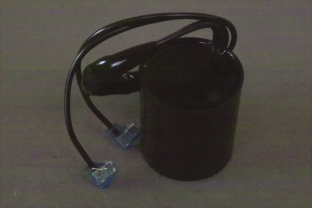 Schwarzer zylindrischer Brötje Kondensator mit zwei angeschlossenen Kabeln in blauer Verbindungstechnik. Der Hintergrund ist neutral grau.