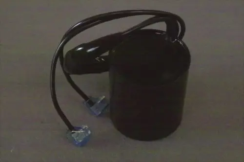 Schwarzer zylindrischer Brötje Kondensator mit zwei angeschlossenen Kabeln in blauer Verbindungstechnik. Der Hintergrund ist neutral grau.