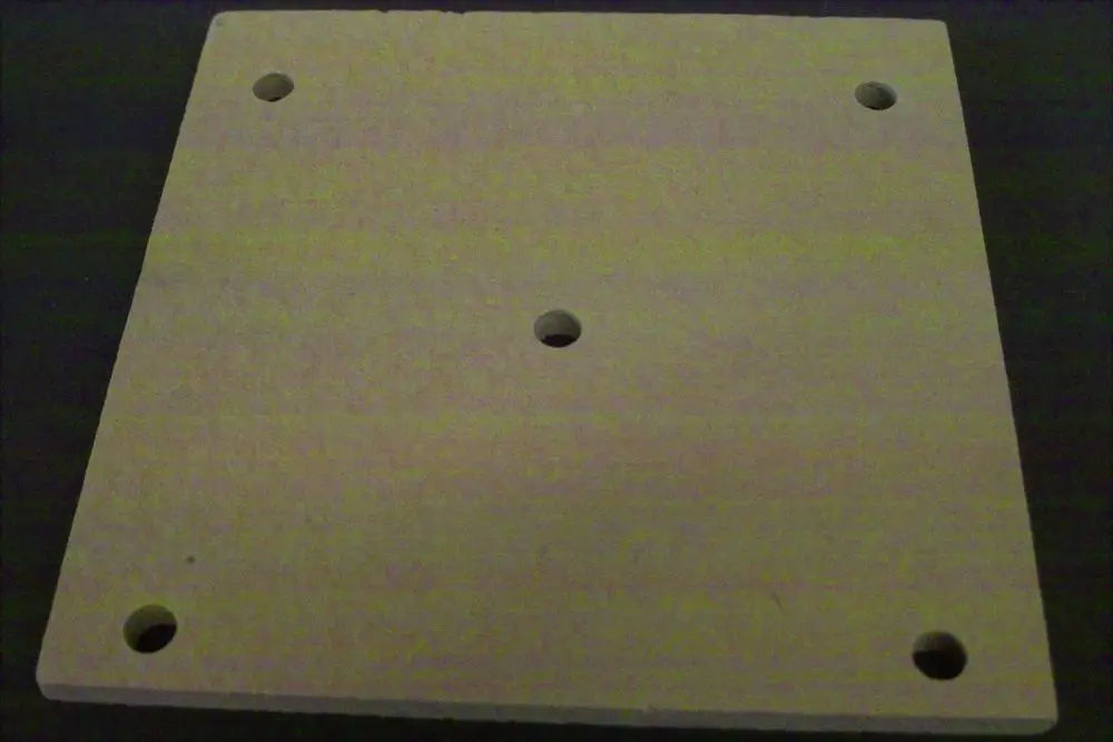 Brötje Kesselisoliermatte aus hellem Material, quadratisch mit mehreren Löchern in den Ecken und einem zentralen Loch, auf dunklem Untergrund abgebildet.