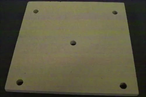 Brötje Kesselisoliermatte aus hellem Material, quadratisch mit mehreren Löchern in den Ecken und einem zentralen Loch, auf dunklem Untergrund abgebildet.