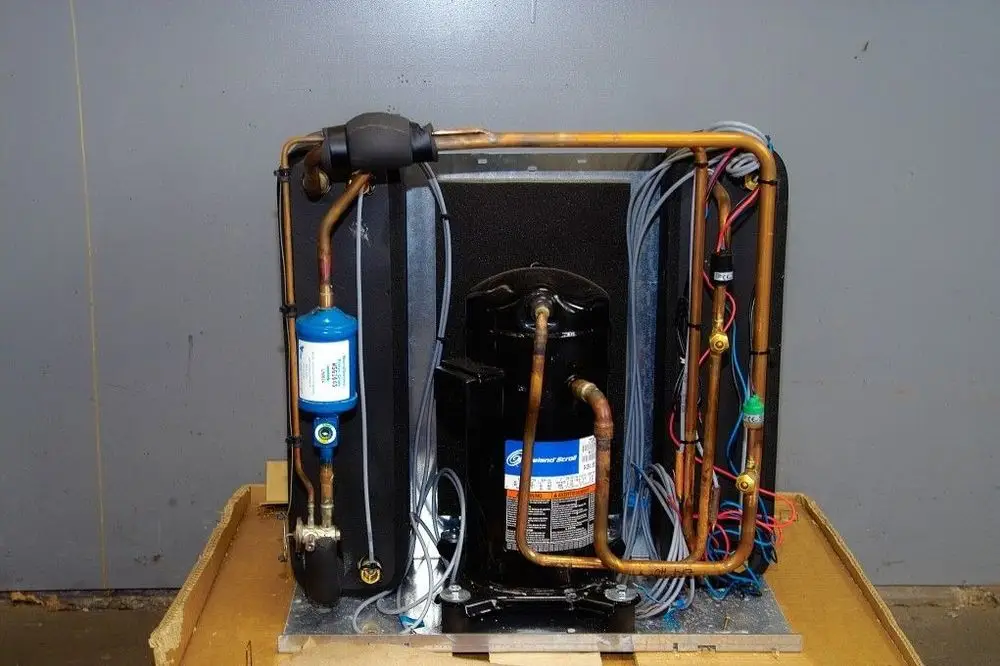 Brötje Kälteteil mit einer Kompressorbaugruppe, Kupferrohren und elektrischen Anschlüssen, montiert auf einer Metallplatte vor neutralem Hintergrund.