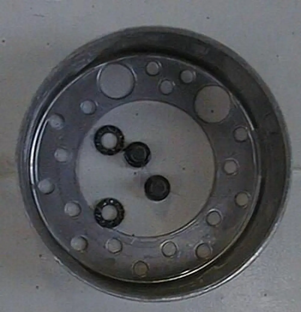 Metalldisk mit mehreren Durchbrüchen, in der Mitte sind drei schwarze Ringe platziert. Produkt möglicherweise von Brötje, Funktion unklar.