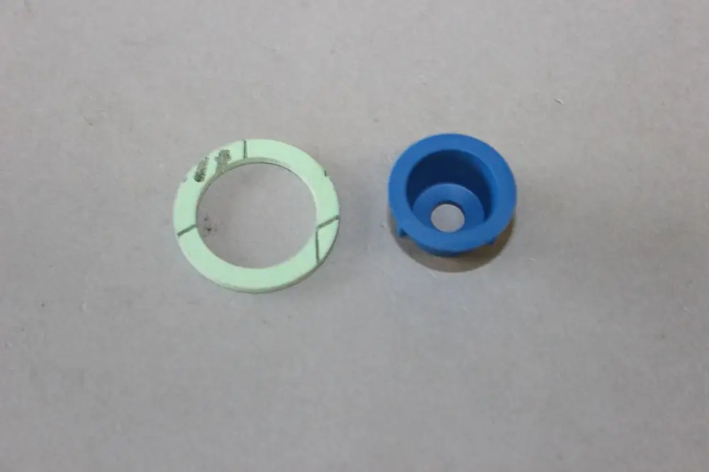 Zwei Kunststoffteile auf grauem Untergrund. Ein grünes, ringförmiges Element mit Linien und ein blaues, hohles Bauteil, wahrscheinlich von Brötje.