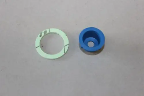 Zwei Kunststoffteile auf grauem Untergrund. Ein grünes, ringförmiges Element mit Linien und ein blaues, hohles Bauteil, wahrscheinlich von Brötje.