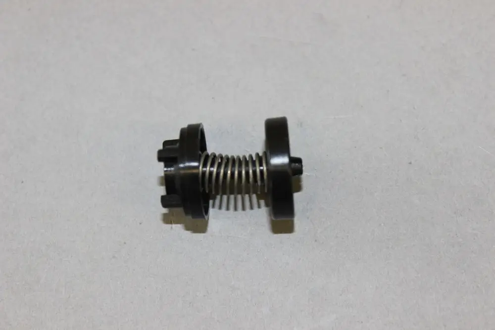 Black spring-loaded component with circular ends, likely part of a Brötje heating system, positioned on a light gray surface.
