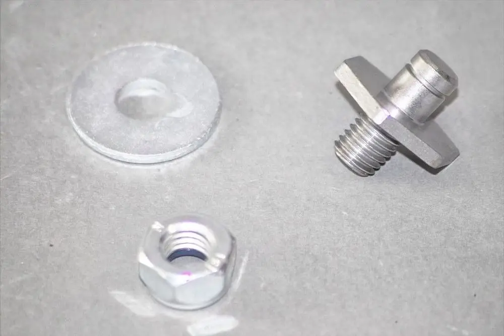 Brötje Zubehörteile, bestehend aus einer runden Unterlegscheibe, einem Gewindebolzen mit schräger Kante und einer sechseckigen Mutter auf grauem Untergrund.