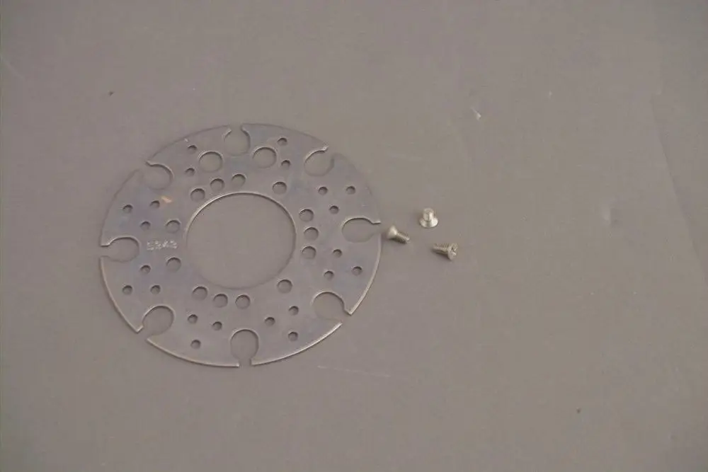 Eine runde Metallplatte mit mehreren Löchern und Aussparungen sowie drei Schrauben, möglicherweise Teil eines Brötje Heizungszubehörs.
