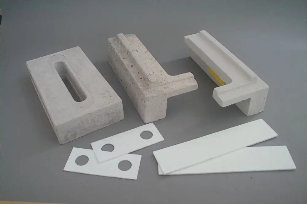 Brötje Ersatzsteine in verschiedenen Formen aus Beton, zusammen mit einigen weißen Platten und Unterlegscheiben, auf grauem Hintergrund angeordnet.