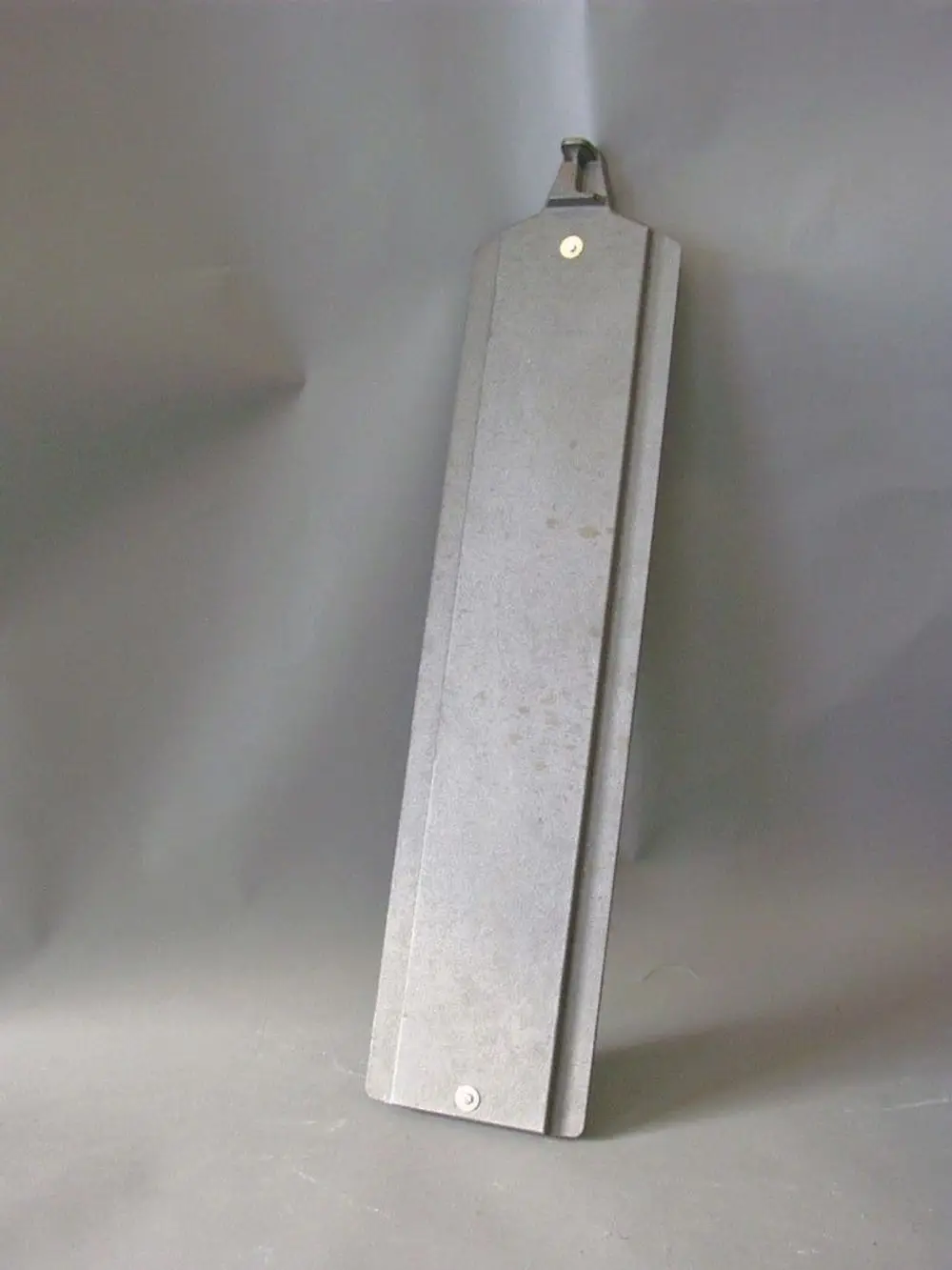 Brötje Wirbulator, flache graue Metallplatte mit einem Haken oben und einer Befestigungsschraube unten, auf grauem Hintergrund stehend.