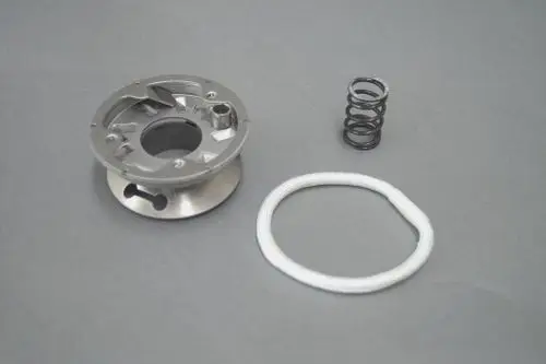 Brötje Umbau-Set bestehend aus einem metallenen Bauteil mit Öffnungen, einer Spiralfeder und einem weißen Dichtungsring, auf grauem Hintergrund angeordnet.