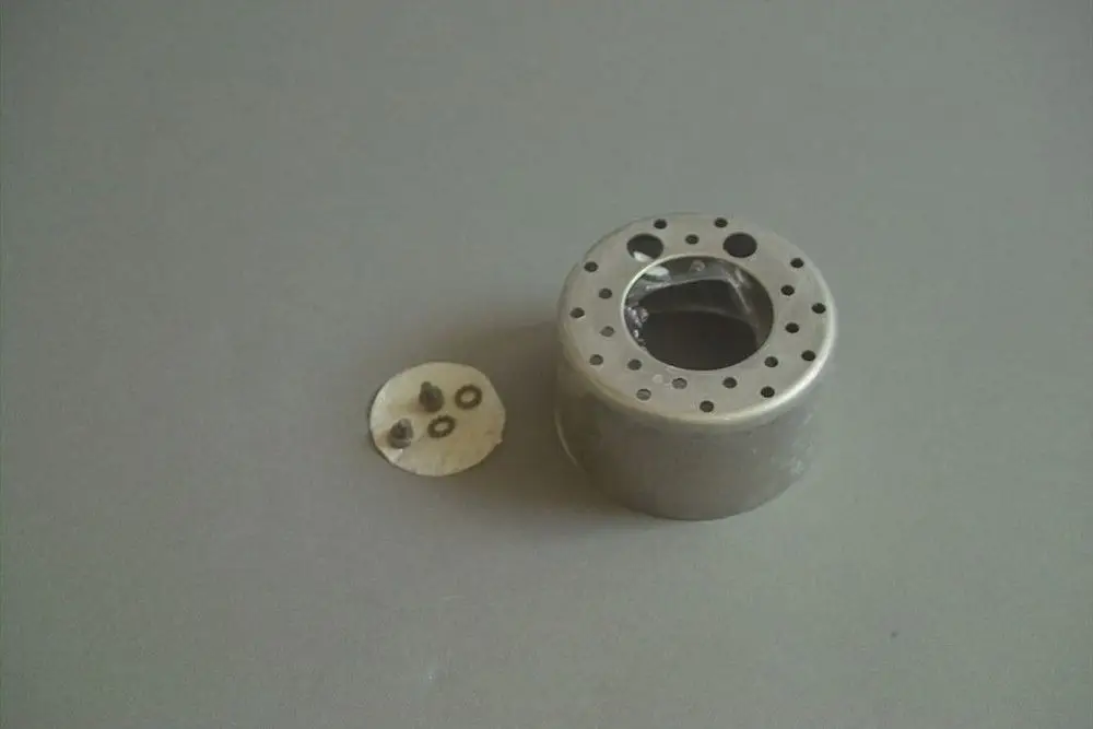 Aluminiumkomponente mit mehreren Löchern und einem offenen Zentrum, daneben eine flache, runde Platte mit drei kleinen Metallteilen. Produkt von Brötje.