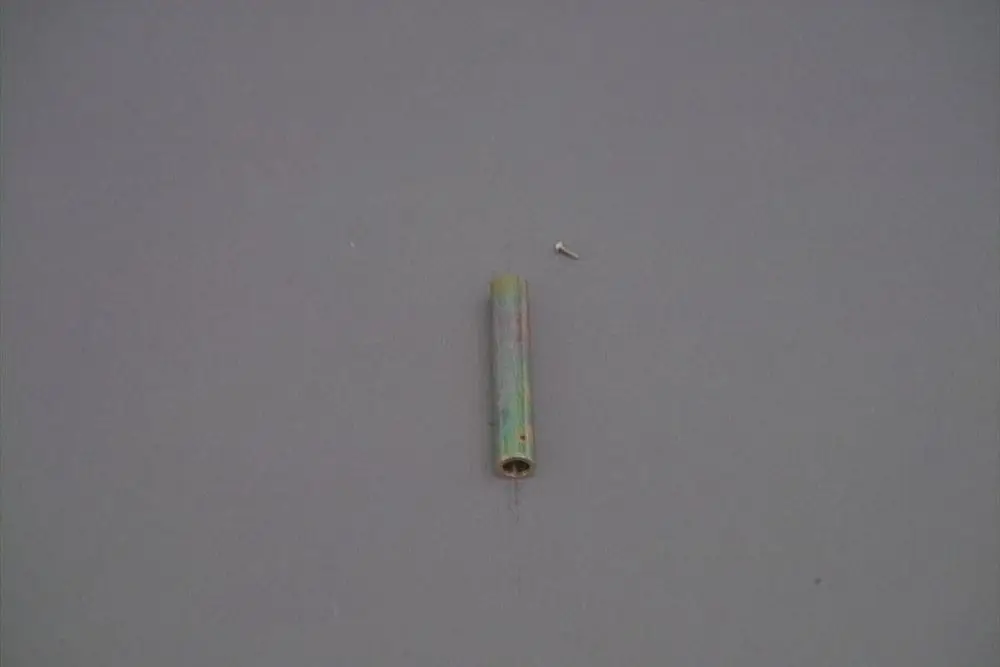 Röhrenförmiges Metallteil in bunten Farben, möglicherweise von Brötje, liegt auf grauem Hintergrund. Daneben befindet sich ein kleiner Schraubenkopf.