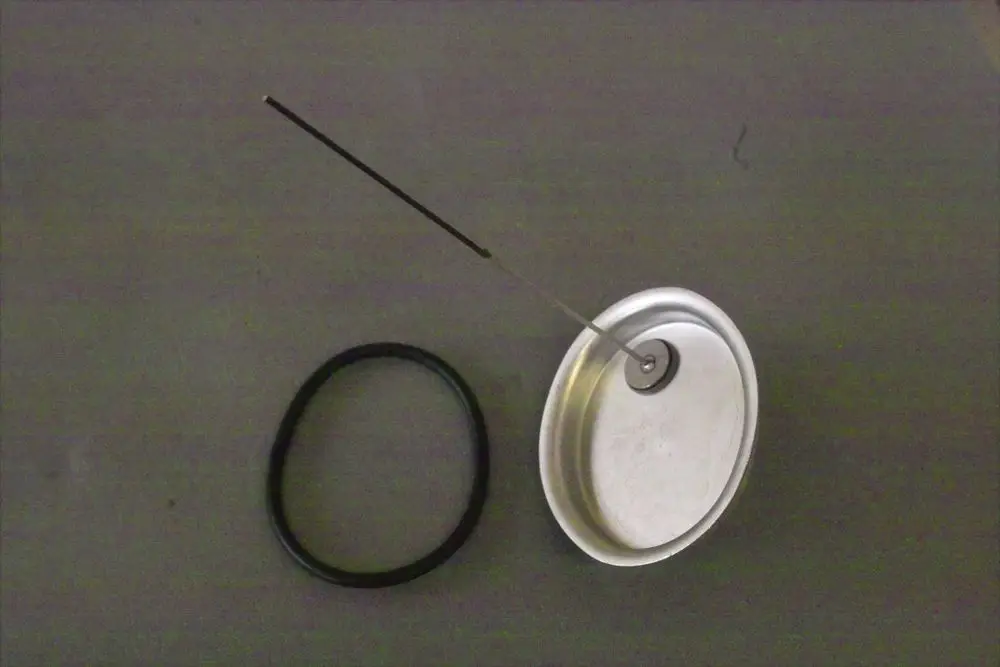 Brötje Handlochdeckel mit einem dünnen Metallstab und einem gummierten O-Ring, auf einer grauen Fläche angeordnet. Der Deckel hat eine zentrale Öffnung und eine glatte Oberfläche.