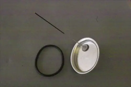 Brötje Handlochdeckel mit einem dünnen Metallstab und einem gummierten O-Ring, auf einer grauen Fläche angeordnet. Der Deckel hat eine zentrale Öffnung und eine glatte Oberfläche.