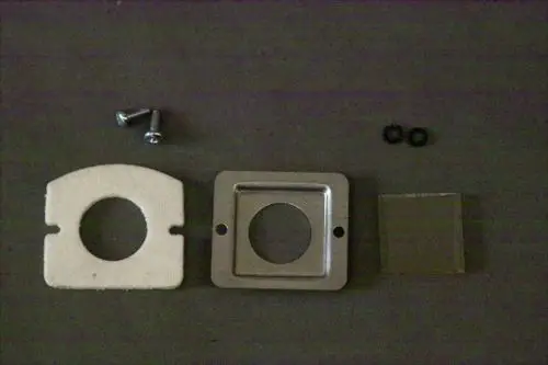 Brötje Schauglas-Set mit einer runden Dichtung, quadratischem Metallrahmen, einer transparenten Abdeckung und zwei Schrauben, alles auf einem dunklen Untergrund.