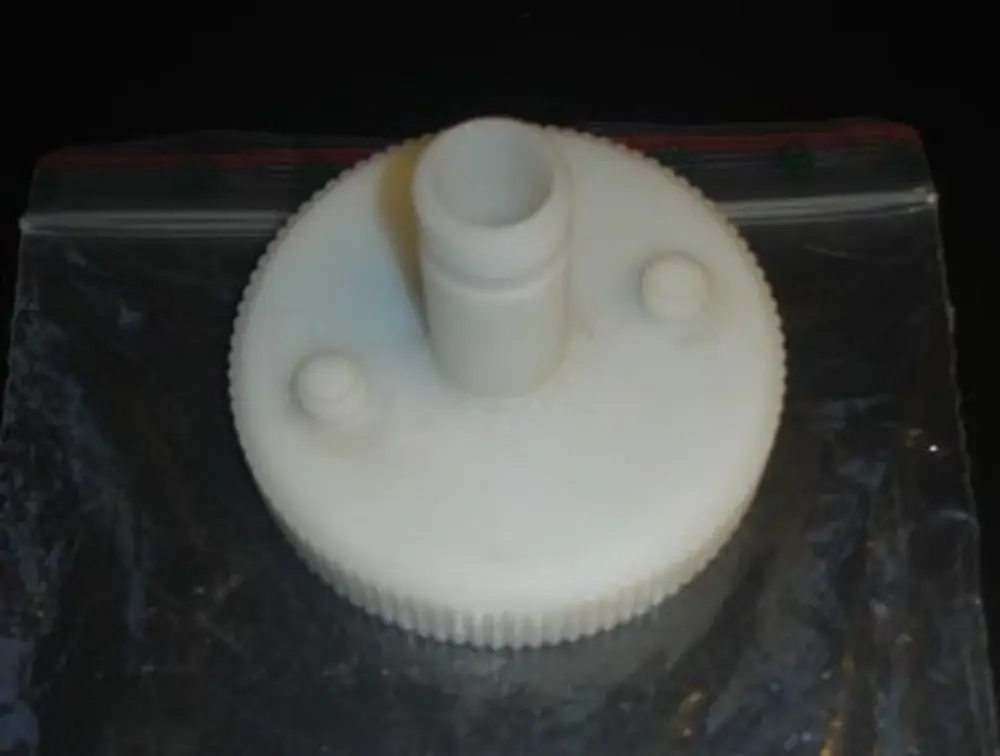 Weißer Kunststoffdeckel mit Messöffnung und einem zentralen Stecker. Der Deckel hat eine gezähnte Kante und ist mit zwei kleinen Erhöhungen versehen. Produkt von Brötje.