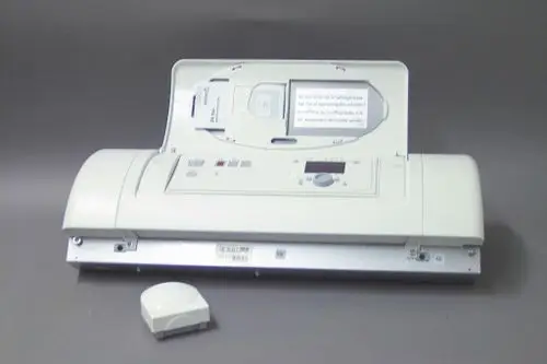 Brötje Heizungsregler in weißer Farbe, geöffnet, mit Bedienfeld und digitalem Display. Separat ein kleiner Sensor erkennbar. Hintergrund grau.