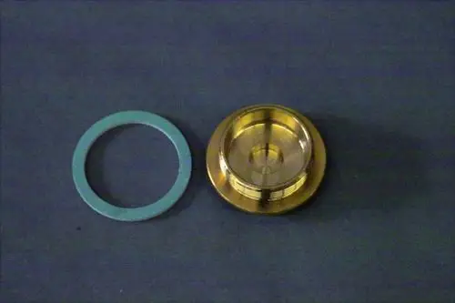 Brötje Verschlussstopfen aus Metall, neben einer grünen Dichtung, auf einem grauen Untergrund. Der Stopfen zeigt ein gewindeteil und eine glatte Oberfläche.