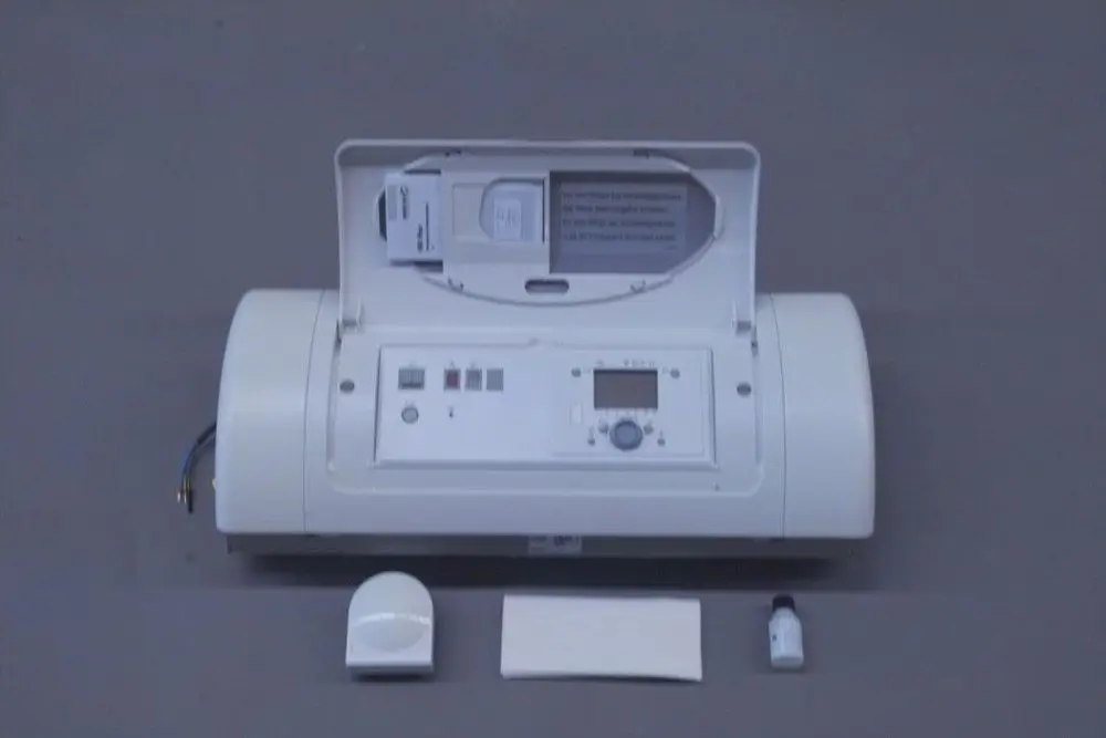 Brötje Regelung mit digitalem Display und mehreren Tasten, dazu Sensor und kleines Fläschchen, auf einem grauen Untergrund angeordnet.