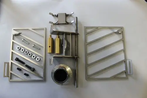 Brötje Ascheaustragung, bestehend aus Metallkomponenten, Bolzen, Schrauben und einer runden Öffnung. Teile angeordnet auf einer hellen Oberfläche.