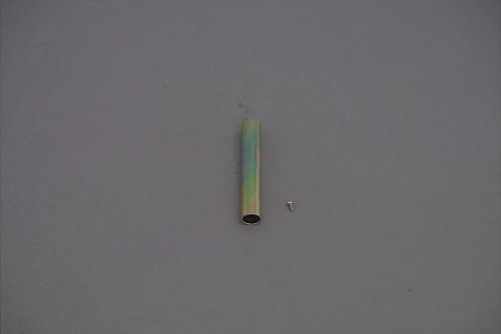 Brötje Röhrchen in zylindrischer Form, metallisch mit einer bunten Oberflächenbehandlung, daneben eine kleine Schraube auf grauem Hintergrund.