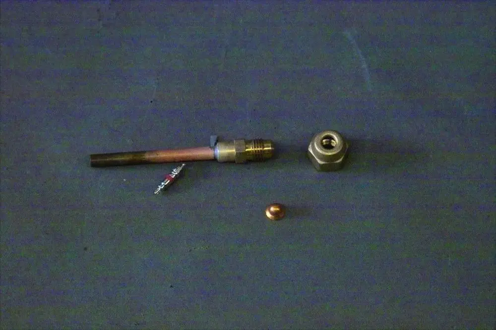 Brötje Schraderverschraubung in verschiedenen metallischen Teilen, darunter ein kurzer Metallrohrabschnitt, eine Mutter und eine kupferne Kugel, auf grauem Untergrund.