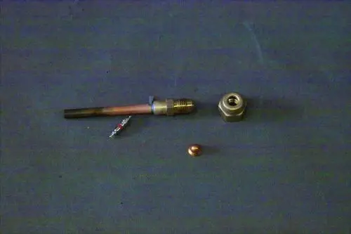 Brötje Schraderverschraubung in verschiedenen metallischen Teilen, darunter ein kurzer Metallrohrabschnitt, eine Mutter und eine kupferne Kugel, auf grauem Untergrund.