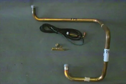 Brötje Sauggasleitung, bestehend aus gebogenen Metallrohren, einem Kabel und einem kleinen eckigen Bauteil. Komponenten sind auf einem grauen Untergrund angeordnet.