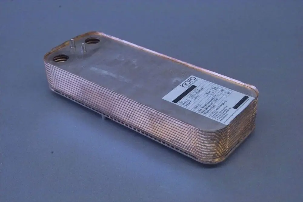 Brötje Wärmetauscher, längliche Metallstruktur mit mehreren Lamellen und Anschlüssen, auf grauem Untergrund. Auf der Oberseite befindet sich ein etikettenähnliches Element.