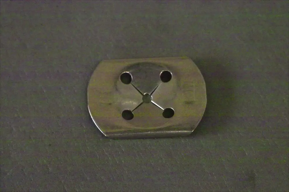 Brötje Halteklammer aus Metall mit vier Bohrungen und einer runden Aussparung in der Mitte, auf grauem Hintergrund abgebildet.