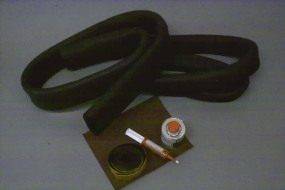 Schwarze, gewundene Schläuche, eine Dose mit rotem Deckel, ein kleiner Behälter mit farbiger Flüssigkeit und ein Stift auf braunem Untergrund. Brötje Reparatur-Set für Solekreise.