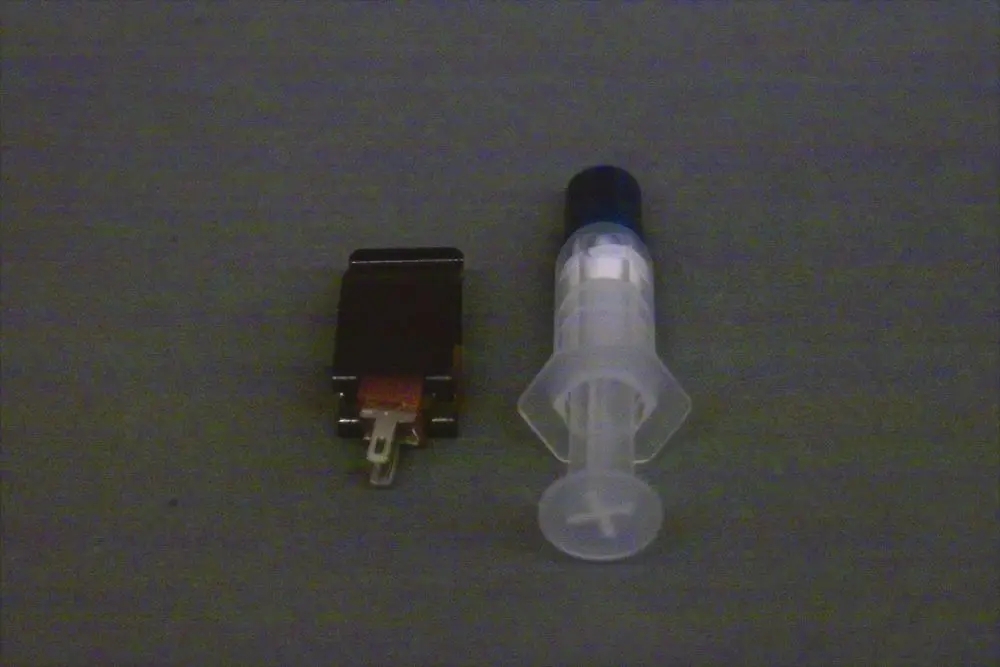 Brötje Vorlauffühler in Form einer kleinen elektronischen Einheit neben einer transparenten Spritze. Beide Objekte auf grauem Hintergrund abgebildet.