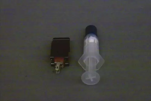 Brötje Vorlauffühler in Form einer kleinen elektronischen Einheit neben einer transparenten Spritze. Beide Objekte auf grauem Hintergrund abgebildet.