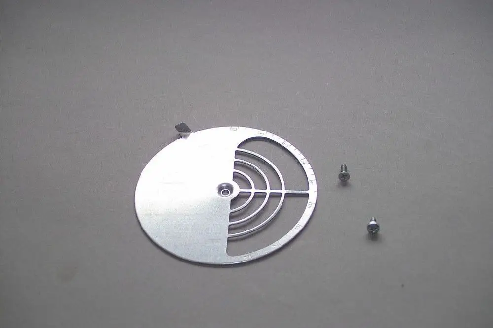 Brötje Luftklappe aus Metall mit kreisförmigem Design und mehreren Öffnungen, zwei kleine Schrauben daneben, auf grauem Untergrund.