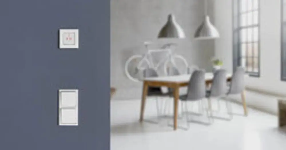 Zwei schlichte Lichtschalter an einer grauen Wand, im Hintergrund ein Essbereich mit einem Holz-Tisch und Stühlen, sowie ein Fahrrad an der Wand. ZigBee Produkte von Danfoss sind möglich.
