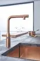 Küchenarmatur in warmem Kupferton von Grohe mit schwenkbarem Auslauf und integriertem Wasserhahn, montiert auf einer Arbeitsplatte aus Beton.