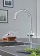 Moderner Chrom-Wasserhahn von Grohe mit geschwungenem Auslauf und integrierter Steuerung, platziert auf einer dunklen Arbeitsfläche neben Teekannen.