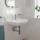 Weißes Waschbecken mit chromfarbener Grohe-Armatur, Wandmontage, Spiegel darüber, Handtuchhalter, Fenster mit Pflanzen im Hintergrund, helle Fliesen.
