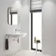 Badezimmeransicht mit wandmontiertem Waschbecken, Spiegel und Grohe-Wasserhahn. Handtuchhalter und kleiner Tisch mit Seife und Handtüchern an Fenster. Helle Farbgebung und Holzfußboden.