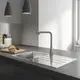 Küchenszene mit Edelstahlspüle, Grohe-Armatur, Holzbrett mit Brot und Utensilien auf einer dunklen Arbeitsfläche. Helle Wände und Pendelleuchten im Hintergrund.