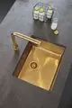 Goldfarbener Küchenwaschtisch mit Grohe-Armatur und abgerundetem Auslauf, umgeben von einer dunklen Arbeitsfläche. Im Hintergrund eine Drahtbox mit Flaschen.