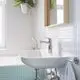 Waschtisch mit Grohe Armatur und runder, weißer Schüssel. An der Wand glänzende Fliesen und ein Holzspiegel, daneben ein Handtuchhalter. Blumentopf mit Pflanze hängt davon.