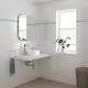 Badezimmeransicht mit einem weißen Waschbecken auf einer Wandplatte, einem verchromten Grohe-Waschtischarmatur, einem Spiegel und einer Pflanze am Fenster.