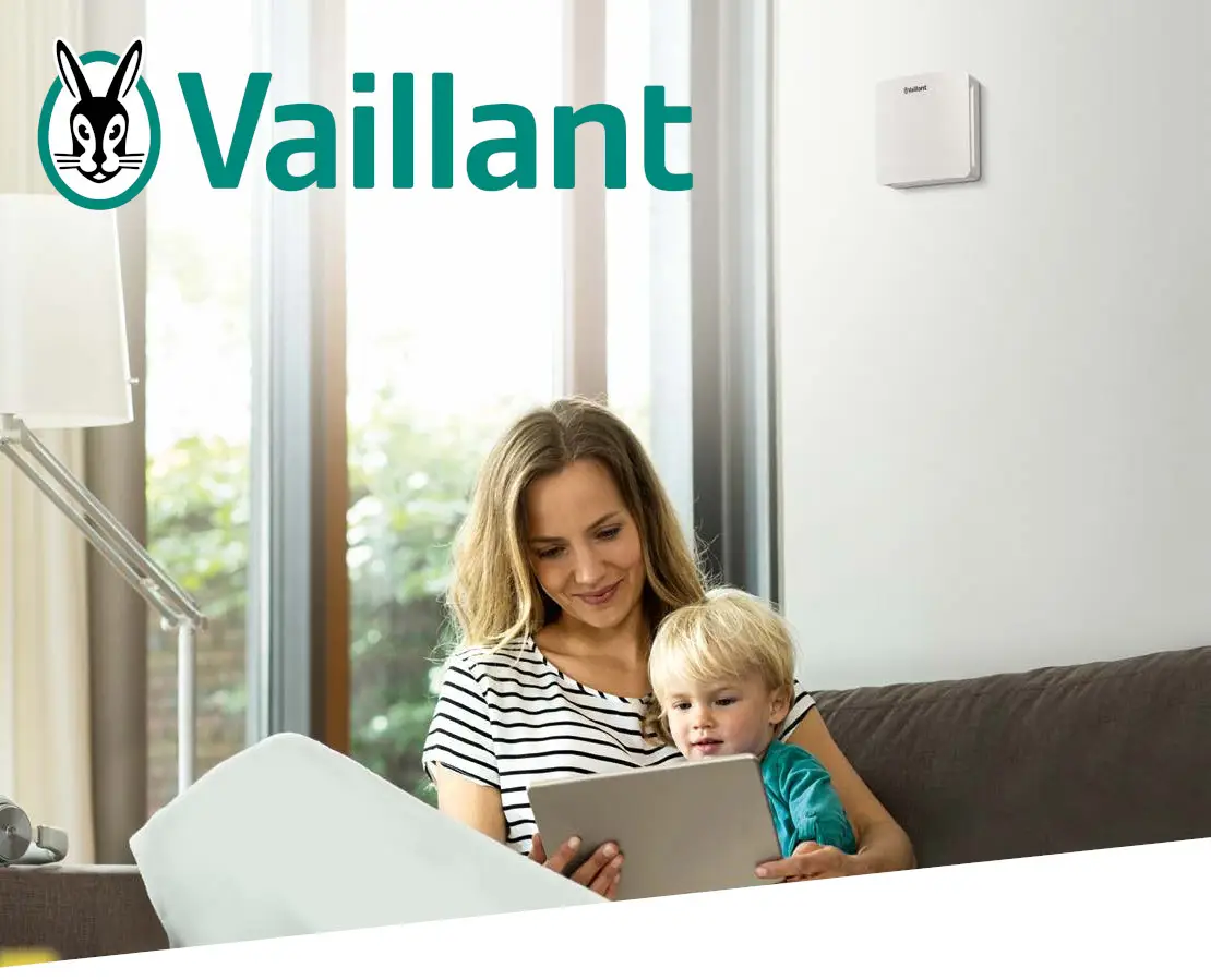 Familie sitzt auf einer Couch in einem hellen Raum. Eine Frau liest auf einem Tablet, während ein kleines Kind neben ihr sitzt. An der Wand ist ein Gerät von Vaillant zu sehen.