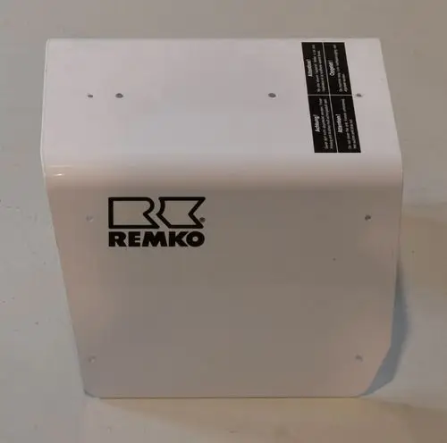 Weißes, rechteckiges Gehäuse mit dem Logo der Marke Remko. Auf der Oberseite befinden sich mehrere Befestigungslöcher und zwei Warnhinweise auf dunklem Hintergrund.